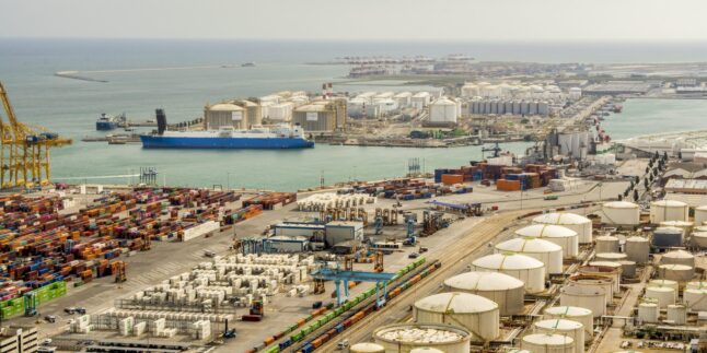 Luftaufnahme eines großen Industriehafens am Mittelmeer mit Tanklagern, Containerterminal und einem Frachtschiff am Kai.