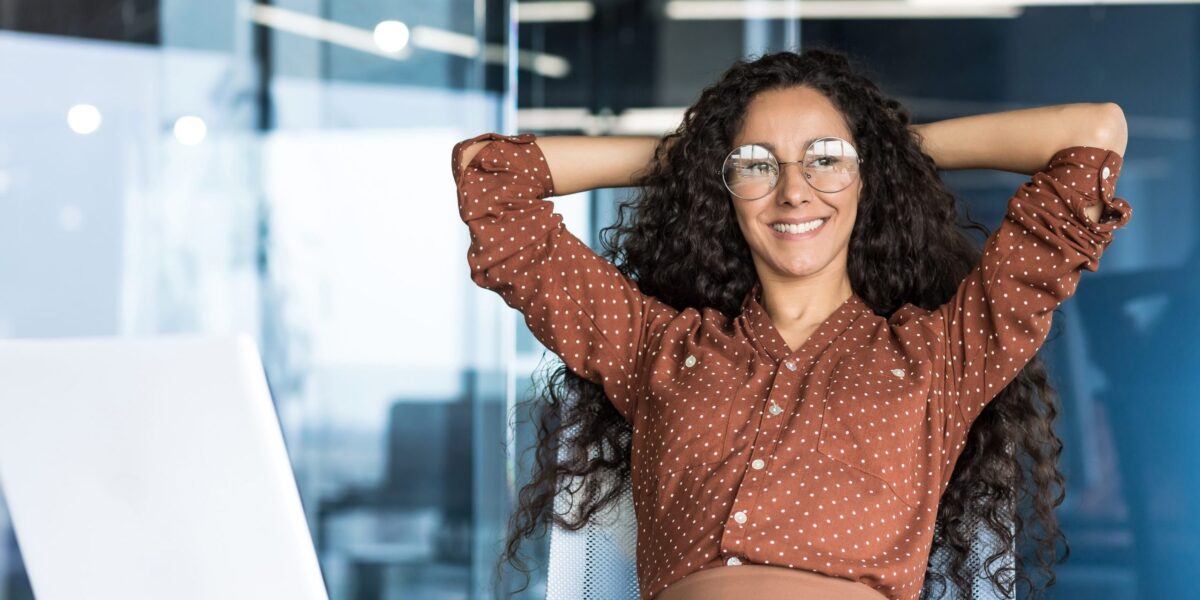 Junge Ingenieurin/Entwicklerin/Programmiererin arbeitet im Büro eines IT-Unternehmens, zufrieden mit ihren Arbeitsergebnissen, macht eine Pause, schaut als Hispa­nic aussehende Frau aus dem Fenster, lächelt, verschränkt die Hände hinter dem Kopf und träumt.