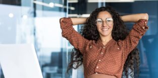 Junge Ingenieurin/Entwicklerin/Programmiererin arbeitet im Büro eines IT-Unternehmens, zufrieden mit ihren Arbeitsergebnissen, macht eine Pause, schaut als Hispa­nic aussehende Frau aus dem Fenster, lächelt, verschränkt die Hände hinter dem Kopf und träumt.