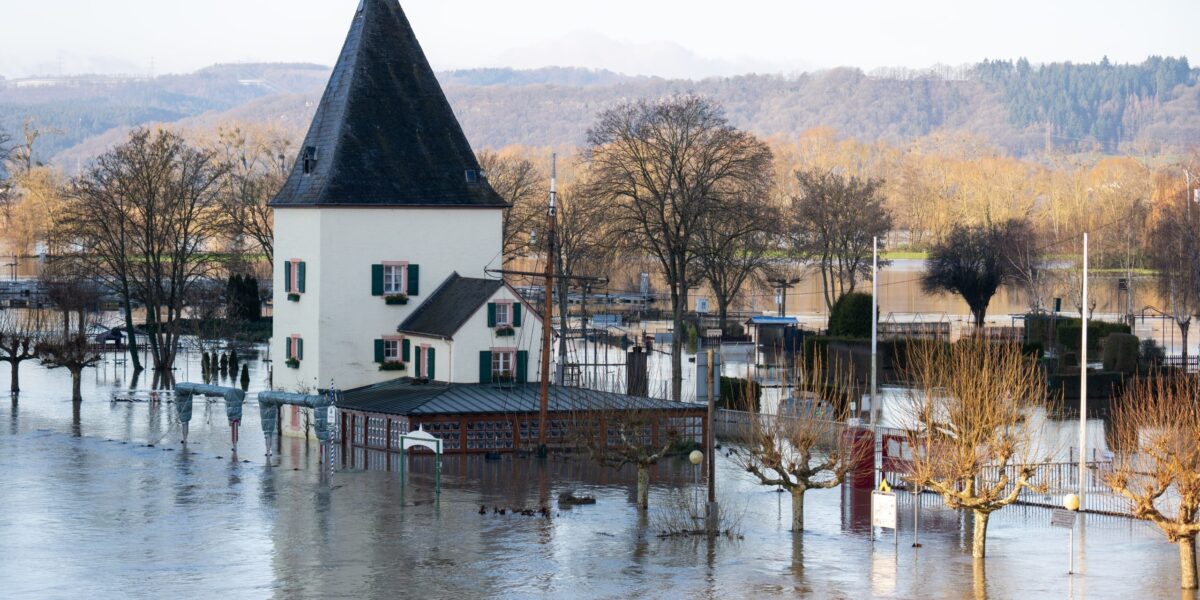 Älteres Gebäude mit Turm am Fluss, umgeben von Bäumen, steht im Hochwasser.