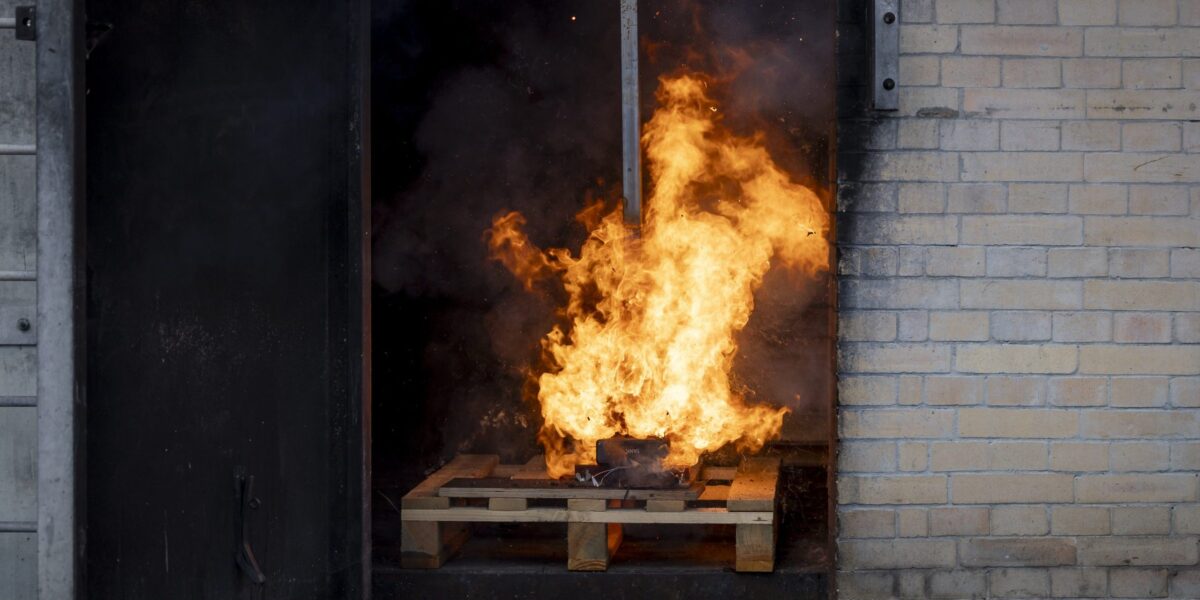 Ein Akku brennt bei einem kontrollierten Brandversuch auf einer Holzpalette. Die Flammen schlagen meterhoch aus dem Gehäuse.
