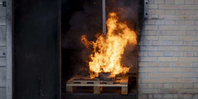 Ein Akku brennt bei einem kontrollierten Brandversuch auf einer Holzpalette. Die Flammen schlagen meterhoch aus dem Gehäuse.