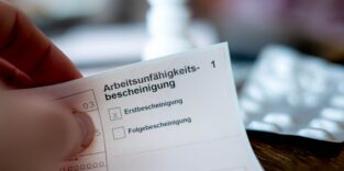 Eine Person hält eine ausgefüllte Arbeitsunfähigkeitsbescheinigung in der Hand. Im Hintergrund ist unscharf ein Tisch mit Medikamenten zu sehen.