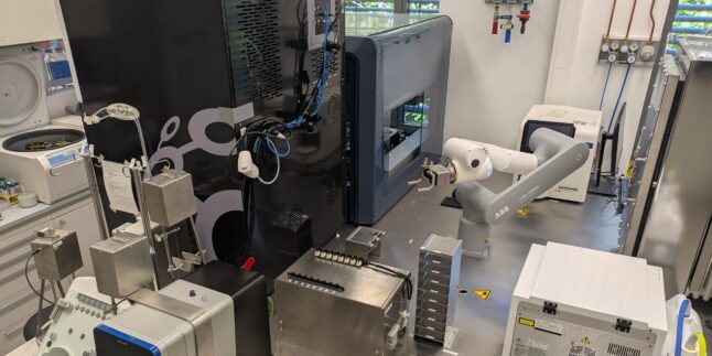 Automatisiertes Labor mit Analysegeräten und Roboterarm, der Proben zwischen den Geräten bewegt.