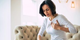 Dunkelhaarige Frau sitzt auf einem beigen Sofa und hält sich mit schmerzverzerrtem Gesicht die Brust – mögliche Symptome eines Herzinfarkts oder einer Herzerkrankung.