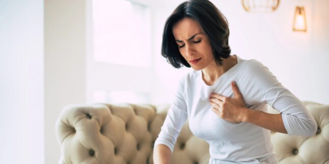 Dunkelhaarige Frau sitzt auf einem beigen Sofa und hält sich mit schmerzverzerrtem Gesicht die Brust – mögliche Symptome eines Herzinfarkts oder einer Herzerkrankung.