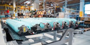 Sieben türkisfarbene Wasserstoff-Hochdruckkammern in einem Metallrahmen auf einem Werkstattgestell, im Hintergrund eine Fertigungsumgebung.