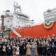 Gruppenfoto vor den Gastankern „Antwerpen" und „Arlon" bei der Taufzeremonie auf der HD Hyundai Heavy Industries Werft in Ulsan am 9. April 2026. Im Hintergrund ist der orangefarbene Rumpf eines der beiden Schiffe zu sehen, davor ein Banner mit der Aufschrift „Naming of Antwerpen & Arlon".