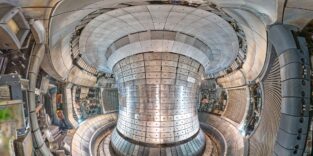 Blick in das Plasmagefäß des Fusionsexperiments ASDEX Upgrade in Garching. Die gesamte Innenwand ist mit metallisch glänzenden Wolfram-Ziegeln ausgekleidet, in der Mitte ragt die zentrale Säule des Tokamaks auf.