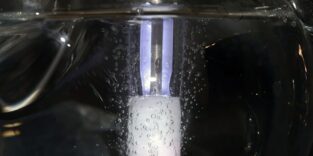 Glasrohr im Wasserbad mit einem weiß schimmernden Katalysator-Block und einem violett leuchtenden Plasmabereich