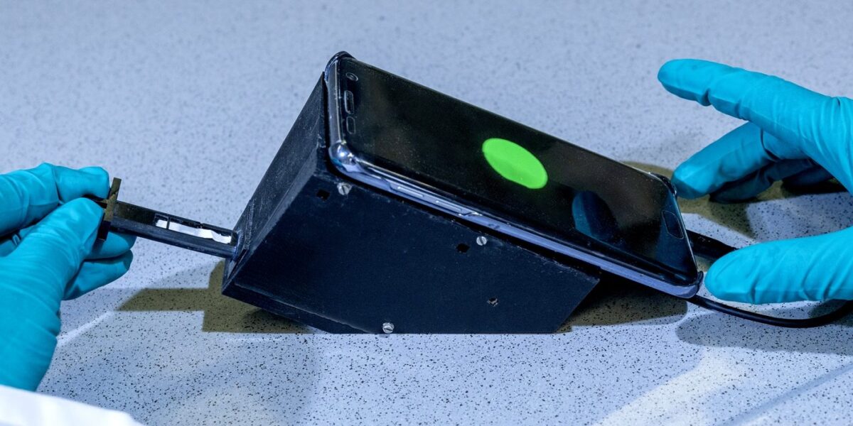 Hände in blauen Laborhandschuhen halten einen schwarzen 3D-gedruckten Aufsatz mit einem Smartphone, auf dessen Display ein grün leuchtender Punkt sichtbar ist – das Fluoreszenzsignal des Wasser-Schnelltests.