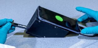 Hände in blauen Laborhandschuhen halten einen schwarzen 3D-gedruckten Aufsatz mit einem Smartphone, auf dessen Display ein grün leuchtender Punkt sichtbar ist – das Fluoreszenzsignal des Wasser-Schnelltests.