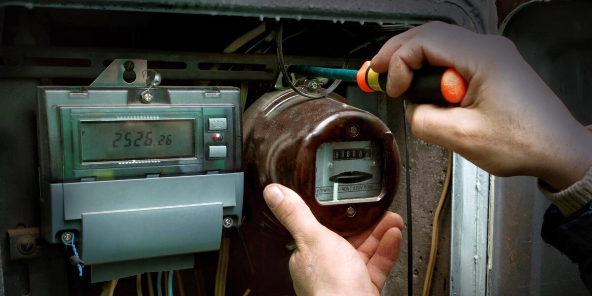 Ein Techniker schraubt mit einem Schraubendreher an einem alten analogen Stromzähler in einem Sicherungskasten. Daneben ist bereits ein digitaler Zähler installiert.