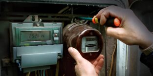Ein Techniker schraubt mit einem Schraubendreher an einem alten analogen Stromzähler in einem Sicherungskasten. Daneben ist bereits ein digitaler Zähler installiert.