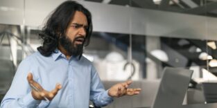 Ein Mann mit langen Haaren und einem blauen Hemd zeigt Frustration und Verwirrung, während er auf den Bildschirm eines Laptops schaut.