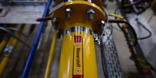 Gelbe Industrierohrleitung mit der Aufschrift „Wasserstoff" und Gefahrgut-Kennzeichnung, verschraubter Flansch, im Hintergrund Anlagentechnik einer Gießerei.