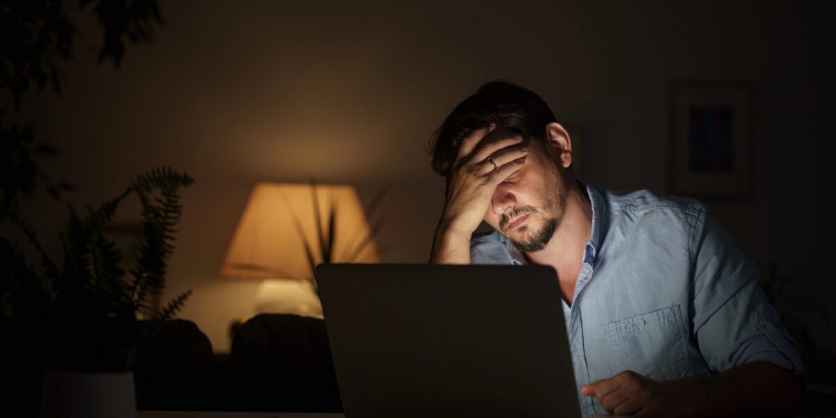 Ein Mann sitzt in einem abgedunkelten Raum an einem Schreibtisch und hält sich den Kopf. Vor ihm steht ein aufgeklappter Laptop.