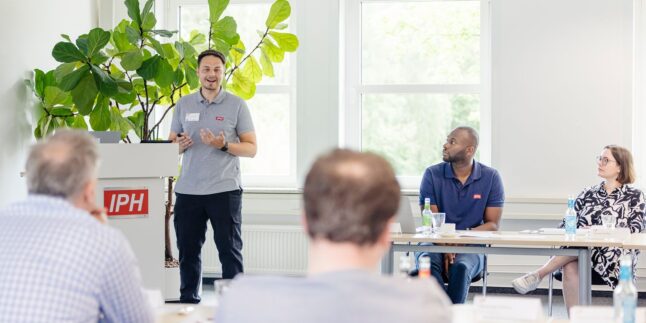 Mann steht in einem Klassenzimmer vor Zuhörenden und erklärt etwas