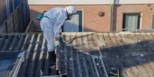 Person in Schutzanzug steht auf Dach und hebt mit Asbest verunreinigte Dachplatte hoch