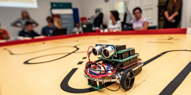 Auf einem gelben Tisch mit schwarzen loopartigen Markierungen sieht man ein kleines Roboterfahrzeug mit Chiplets, schwarzen Rädern und roten Kabeln. Im Hintergrund sieht man verschwommen Menschen.