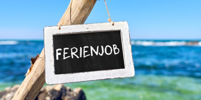 Vor blauem Gewässer sieht man einen Holzpfahl. Daran hängt ein rechteckiges Schild, weiß umrandet mit schwarzem Hintergrund. In weißer Schrift steht dort in Großbuchstaben: Ferienjob.