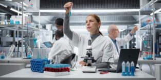 Man sieht eine weibliche Forscherin, sowie weitere Forscher hinter ihr, in einem Labor. Vor ihr auf dem Tisch stehen verschiedene Reagenzgläser mit Proben, ein Mikroskop, sowie ein Laptop. In ihrer Hand hält sie eine Probe.