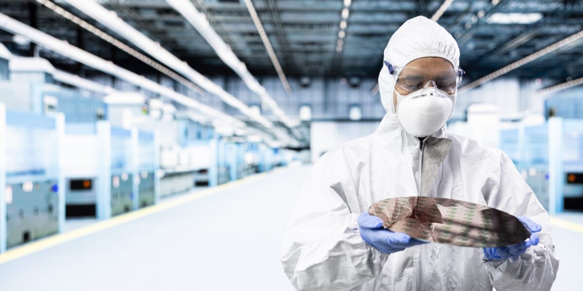 Man sieht einen Mann in einem weißen Ganzkörperschutzanzug mit Maske und Schutzbrille. Er hat blaue Handschuhe an und hält eine Platine. Er steht in einem Labor.