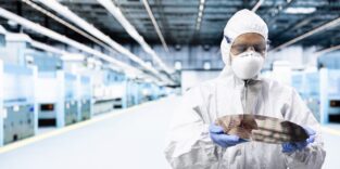 Man sieht einen Mann in einem weißen Ganzkörperschutzanzug mit Maske und Schutzbrille. Er hat blaue Handschuhe an und hält eine Platine. Er steht in einem Labor.