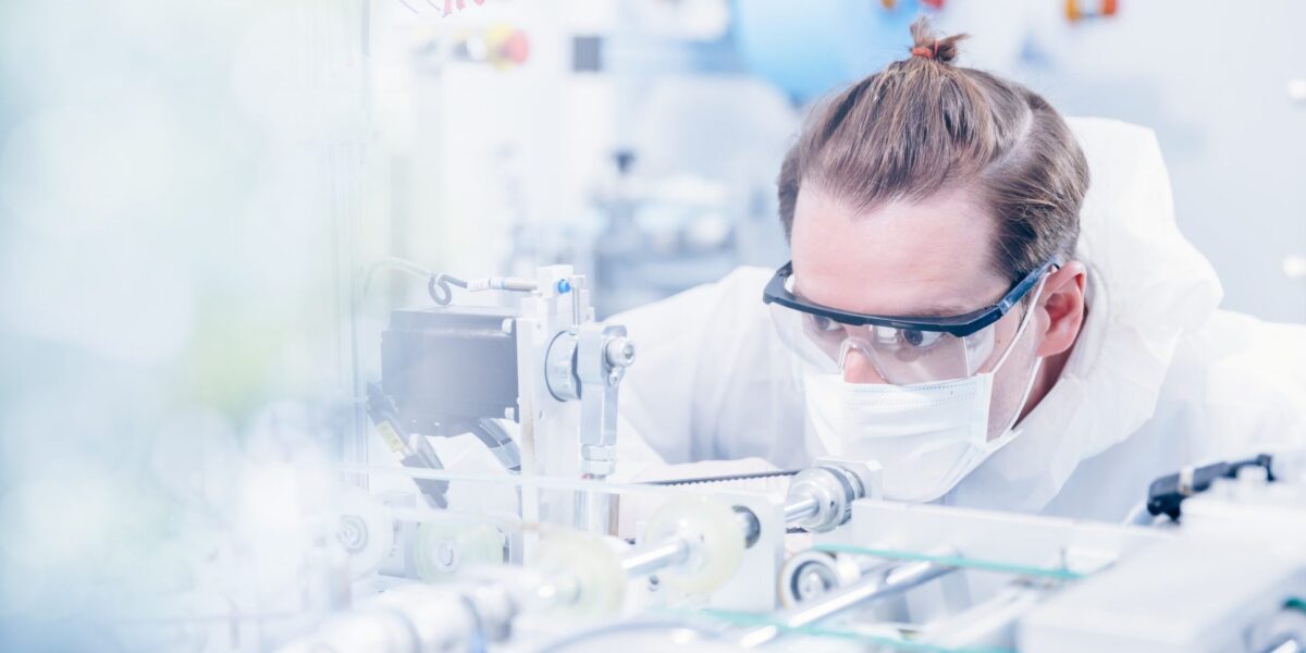 Man sieht einen Mann mit braunen Haaren und einem Dutt, er trägt eine Schutzbrille und ist im Labor. Er trägt einen weißen Schutzanzug und einen weißen Mundschutz.