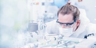 Man sieht einen Mann mit braunen Haaren und einem Dutt, er trägt eine Schutzbrille und ist im Labor. Er trägt einen weißen Schutzanzug und einen weißen Mundschutz.