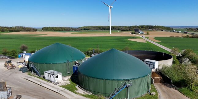 Luftaufnahme einer Biogasanlage mit zwei grünen Fermenterkuppeln, dahinter Felder und eine Windkraftanlage