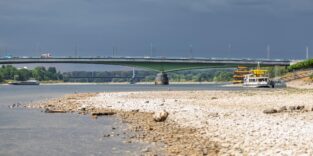 Bis auf die Fahrrinne ausgetrocknetes Flußbett (Rhein), im Hintergrund zwei Brücken mit düsterem Himmel
