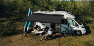 Campervan vor Wald mit ausgefahrener Solarmarkise, zwei Leute an einem Tisch darunter
