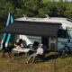 Campervan vor Wald mit ausgefahrener Solarmarkise, zwei Leute an einem Tisch darunter