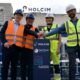 Vier Personen drücken in einem Zementwerk auf einen roten Knopf.