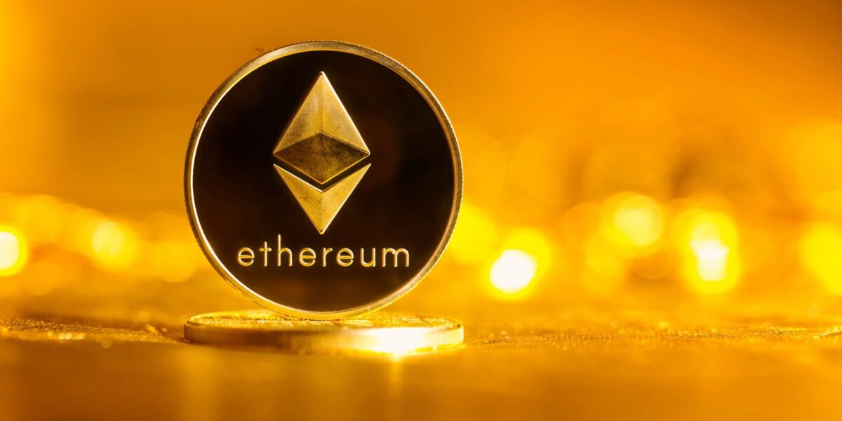 Goldfarbene Münze mit dem Ethereum-Logo und Schriftzug, aufrecht stehend vor unscharfem gelb-orangefarbenem Hintergrund.