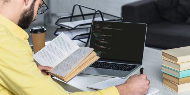 Ein Mann sitzt mit einem Buch am Computer.