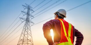 Ein Elektroingenieur steht vor einer Trasse, um zu sehen, wie die Produktion und der Export von Elektrizität an großen Hochspannungsmasten geplant werden können.