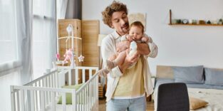 Ein junger Mann mit braunen Haaren und Bart hält ein Baby in den Armen und gibt ihm die Milchflasche. Hinter ihm sind ein weißen Babybett und ein großes Bett zu sehen.