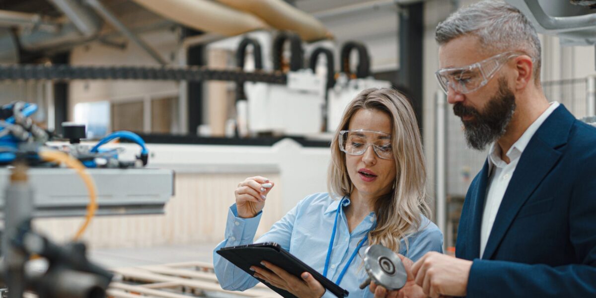 Weibliche Ingenieurin und männlicher Projektmanager stehen in einer modernen Industriehalle neben einem präzisen Roboterarm.