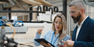 Weibliche Ingenieurin und männlicher Projektmanager stehen in einer modernen Industriehalle neben einem präzisen Roboterarm.
