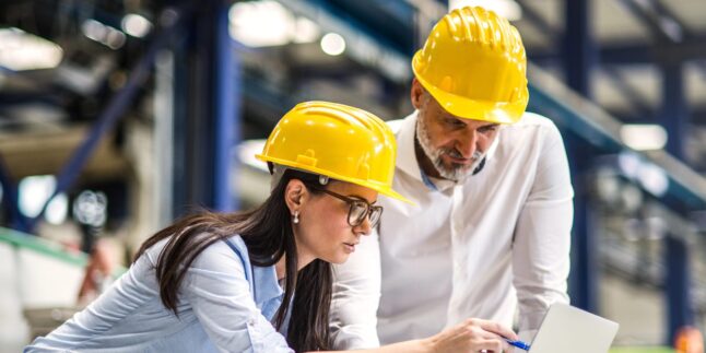 Zwei Wirtschaftsingenieure in einer modernen Industriehalle. Im Hintergrund sind Fertigungsanlagen, Roboterarme und automatisierte Produktionssysteme zu sehen.