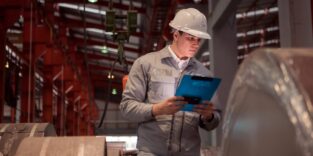 Qualitätsingenieur mit Notizbrett und weißen Schutzhelm inspiziert das Innere eines großen Metallrohrs in einer Fabrik.