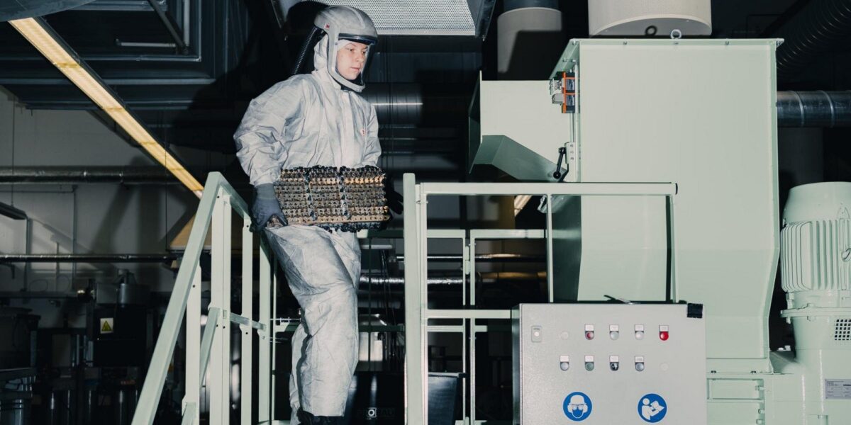 Person in grauem Schutzanzug mit Helm trägt ein Batteriemodul eine Treppe an einer Industrieanlage hinauf