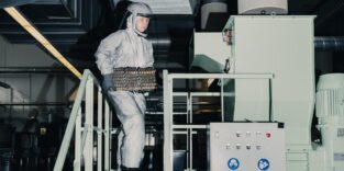Person in grauem Schutzanzug mit Helm trägt ein Batteriemodul eine Treppe an einer Industrieanlage hinauf