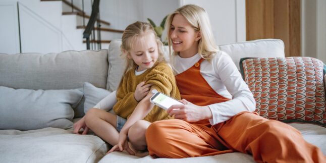 Person sitzt auf einem Bett, am Oberarm ein Glukosesensor, in den Händen ein Smartphone