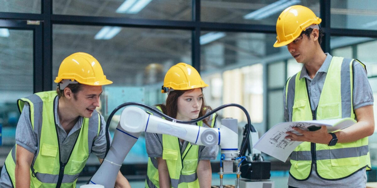 Eine Gruppe junger Praktikanten aus dem Ingenieursberuf steht um einen Roboterarm versammelt.