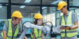 Eine Gruppe junger Praktikanten aus dem Ingenieursberuf steht um einen Roboterarm versammelt.