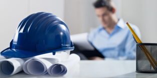 Bauhelm und Pläne im Vordergrund, Mann verschwommen im Hintergrund