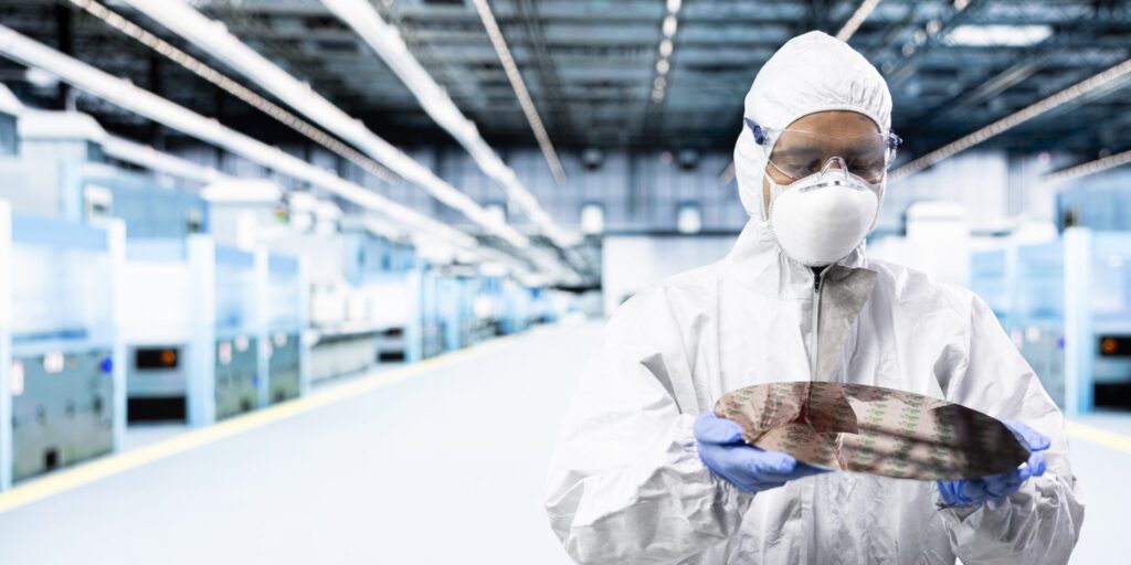 Man sieht einen Mann in einem weißen Ganzkörperschutzanzug mit Maske und Schutzbrille. Er hat blaue Handschuhe an und hält eine Platine. Er steht in einem Labor.